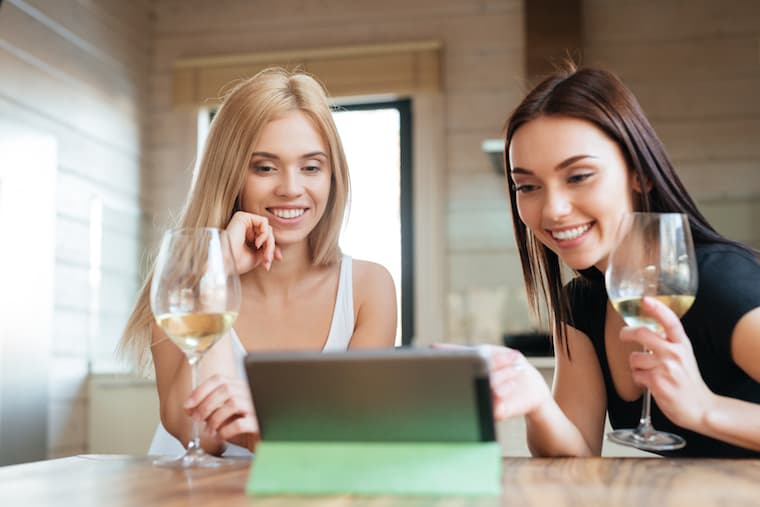 Two smiling friends sitting by the table in kitchen while drinking wine and looking at the tablet computer